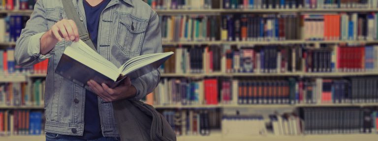 En ung person i jeansjacka håller i en öppen bok, bläddrar i boken. I bakgrunden syns bokhyllorna i ett bibliotek.